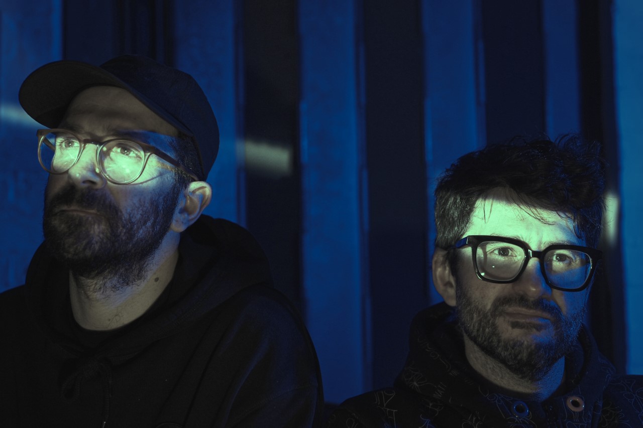 The photo shows the faces of two men in glasses standing in a dark room. On their faces, in the areas of their eyes, a shaft of light pierces.