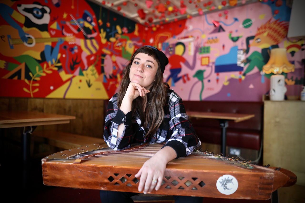 The photo shows a seated woman with a Lithuanian box zither on her lap. It is a stringed, plucked instrument. The woman holds the instrument with one hand and rests it under her chin with the other.
