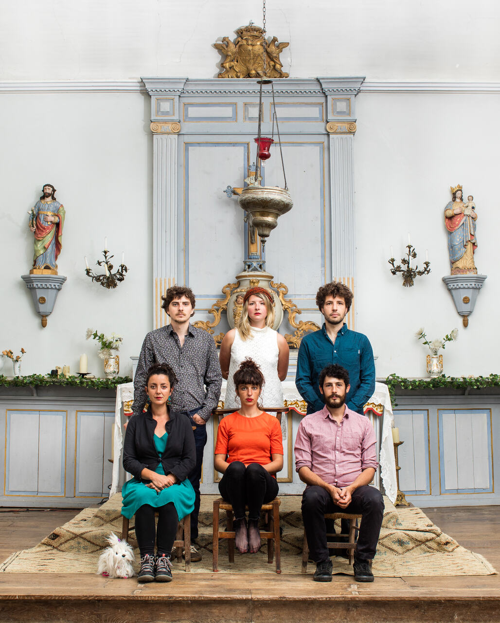 The photo shows six people, women and men, colorfully dressed, in a richly decorated room in the Baroque style. Three are sitting on chairs in one row, another three are standing right behind them.