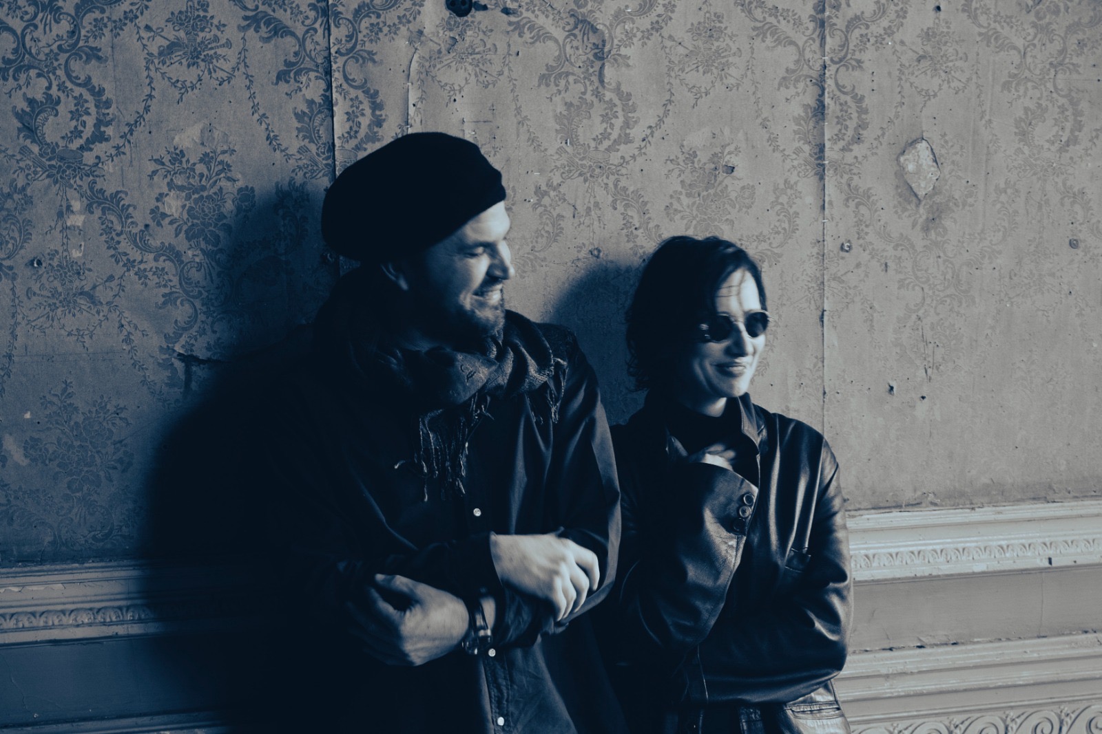 The photo shows a smiling woman and a man leaning against a wall. The man looks askance at the woman.