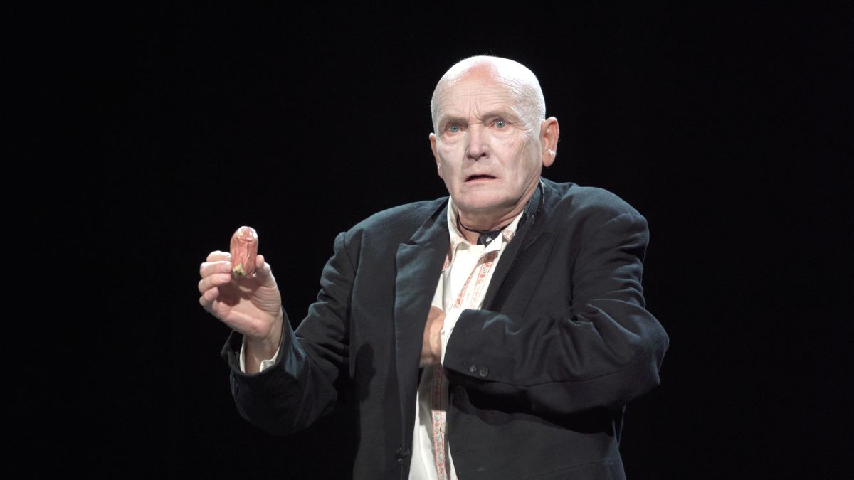 łysy starszy mężczyzna w marynarce, aktor mim, z poważnym wyrazem twarzy sięga po coś do kieszeni, trzymając jakiś przedmiot przypominający nos w drugiej ręce