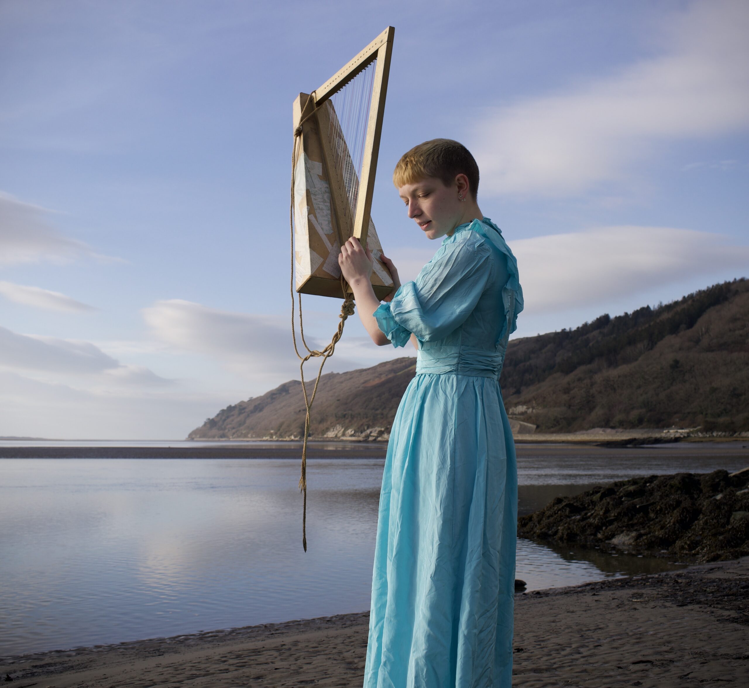 The photo shows a person in a blue dress holding a small harp against the backdrop of mountains and the sea.