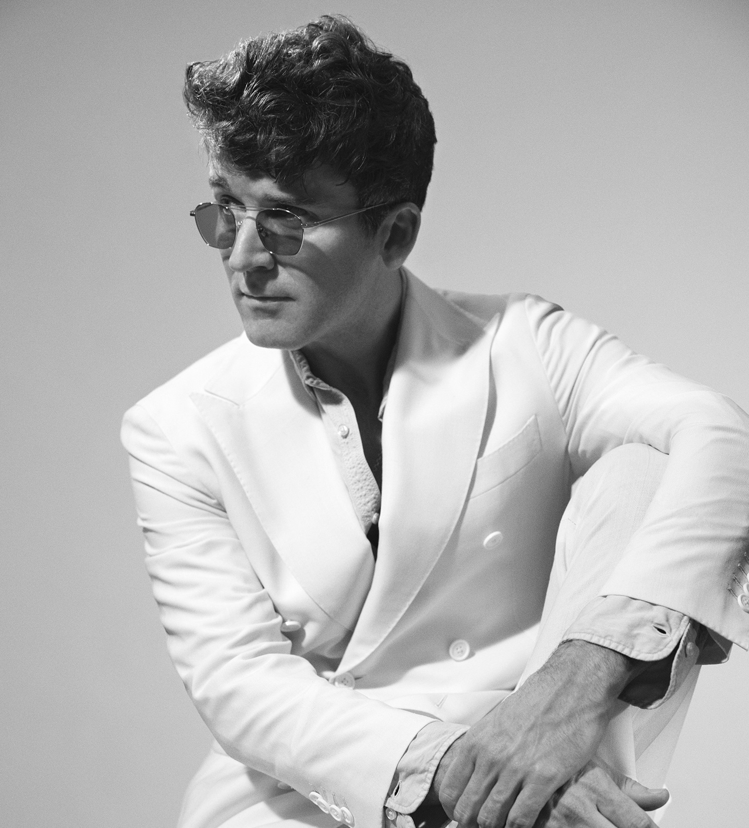 Na zdjęciu widać siedzącego mężczyznę w okularach przeciwsłonecznych w garniturze.