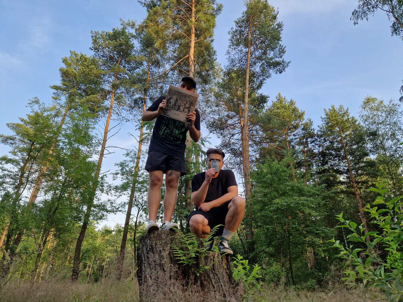 The photo shows two men in the forest. One is crouching, the other standing on a felled tree trunk.