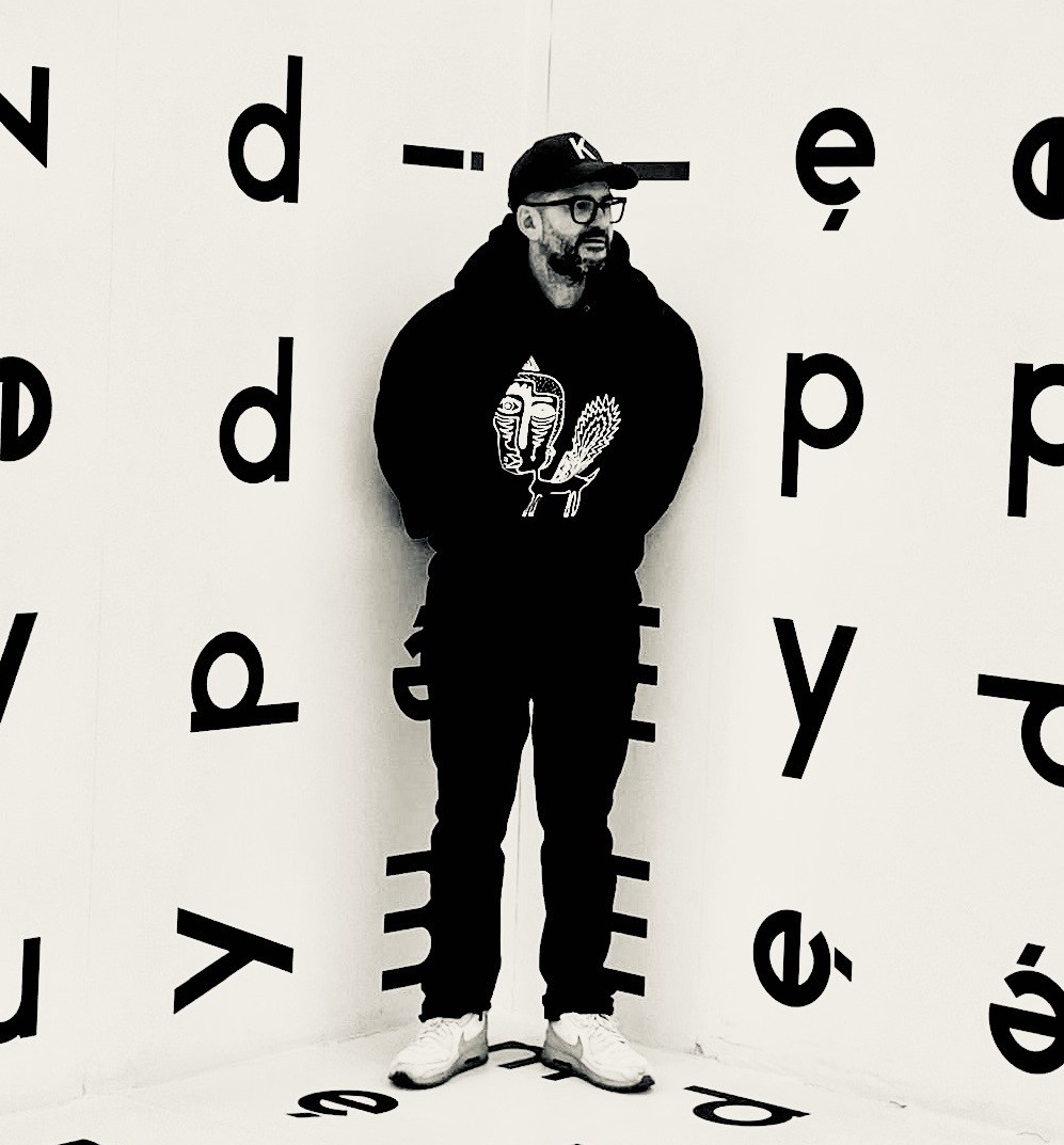 The photo shows a man standing in the corner of a room covered in large letters on the walls.