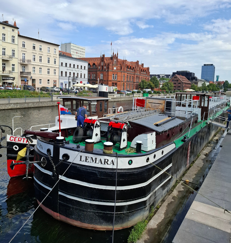 Na pierwszym planie zacumowana przy nabrzeżu barka „Lemara” o czarno-białym kadłubie i zielonym pokładzie. Obok barki znajduje się mniejsza jednostka pływająca, a na pokładzie widoczna jest osoba wykonująca prace przy łodzi. W tle rozciąga się rzeka oraz zabudowa nadbrzeżna, w tym charakterystyczny czerwony ceglany budynek i jasne kamienice. Po prawej stronie widoczny jest deptak biegnący wzdłuż nabrzeża. Nad miastem rozpościera się częściowo zachmurzone niebo z przejaśnieniami.