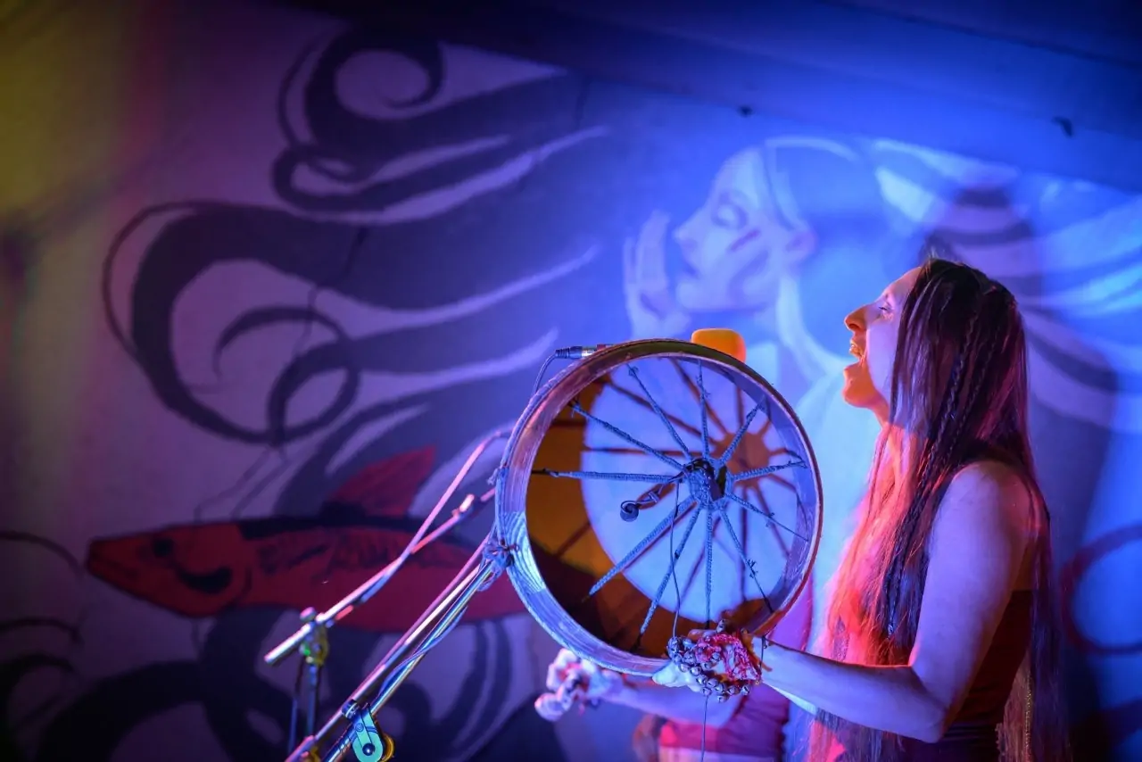 The photo shows the left profile of a young woman singing and playing the hoop drum, against the backdrop of a mural depicting the face of a woman with flowing long black hair.