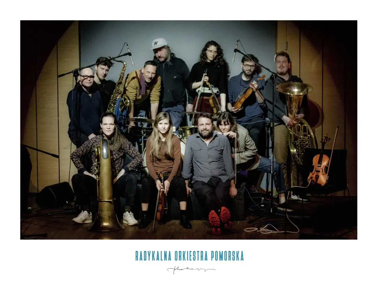 The photo shows eleven people in a recording studio against a wooden wall and a gray square sheet. Seven people are standing, holding instruments in their hands, including tuba, violin, saxophone. Four people are sitting, in front of these seven, two of them with instruments: tuba, violin. There is a violin on the chair to the left. Above them, there are two microphones on stands. Drums are placed between the standing group and the seated group.
