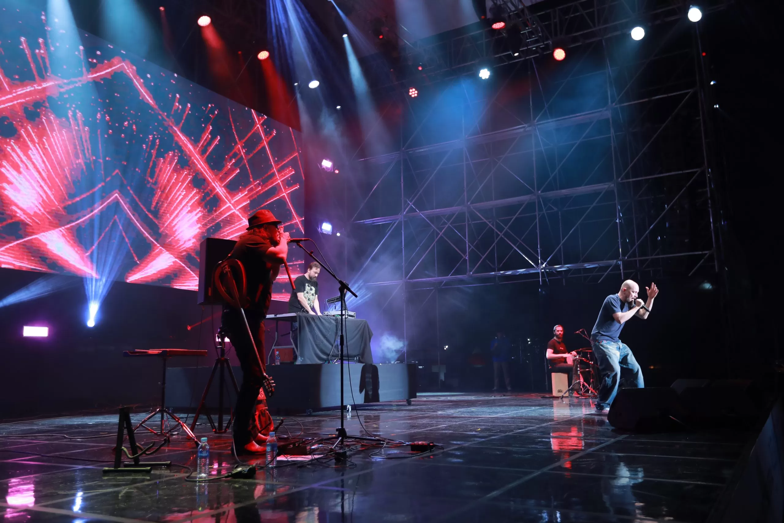 The photo shows four men on a concert stage. The first from the left, he has a guitar slung over his side, he is wearing sunglasses, a hat and he is playing into a microphone that stands in front of him on a stand, on a harp. In the background, right behind, at the DJ table, there is a bearded man in a black T-shirt. On the right side of the photo, a bald man in jeans and a blue T-shirt sings into a microphone. Just behind him in the background, a bearded man is sitting on a cajon and playing the djembe. On the left side of the photo there is a large screen on which red laser shapes are displayed.