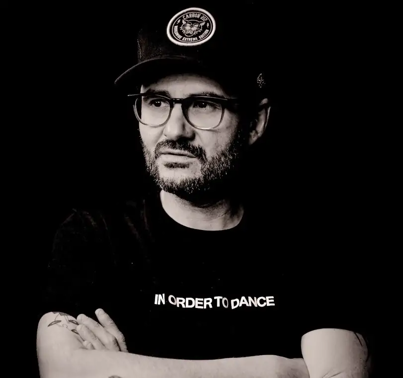 The black and white photo shows a man wearing a baseball cap, glasses and a beard. The man has his arms crossed on his chest.