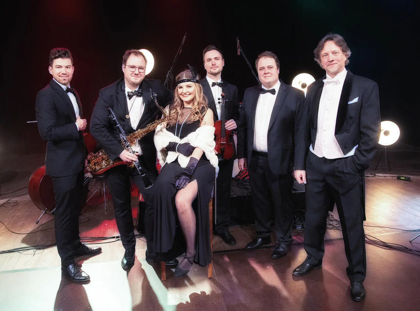 5 mężczyzn we frakach, białych koszulach, na scenie, między nimi kobieta z rozpuszczonymi włosami, w stroju retro.