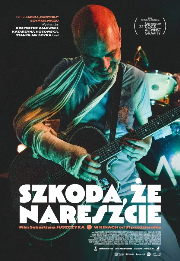łysy mężczyzna z ręką w gipsie na temblaku, oświetlony scenicznie, gra na gitarze