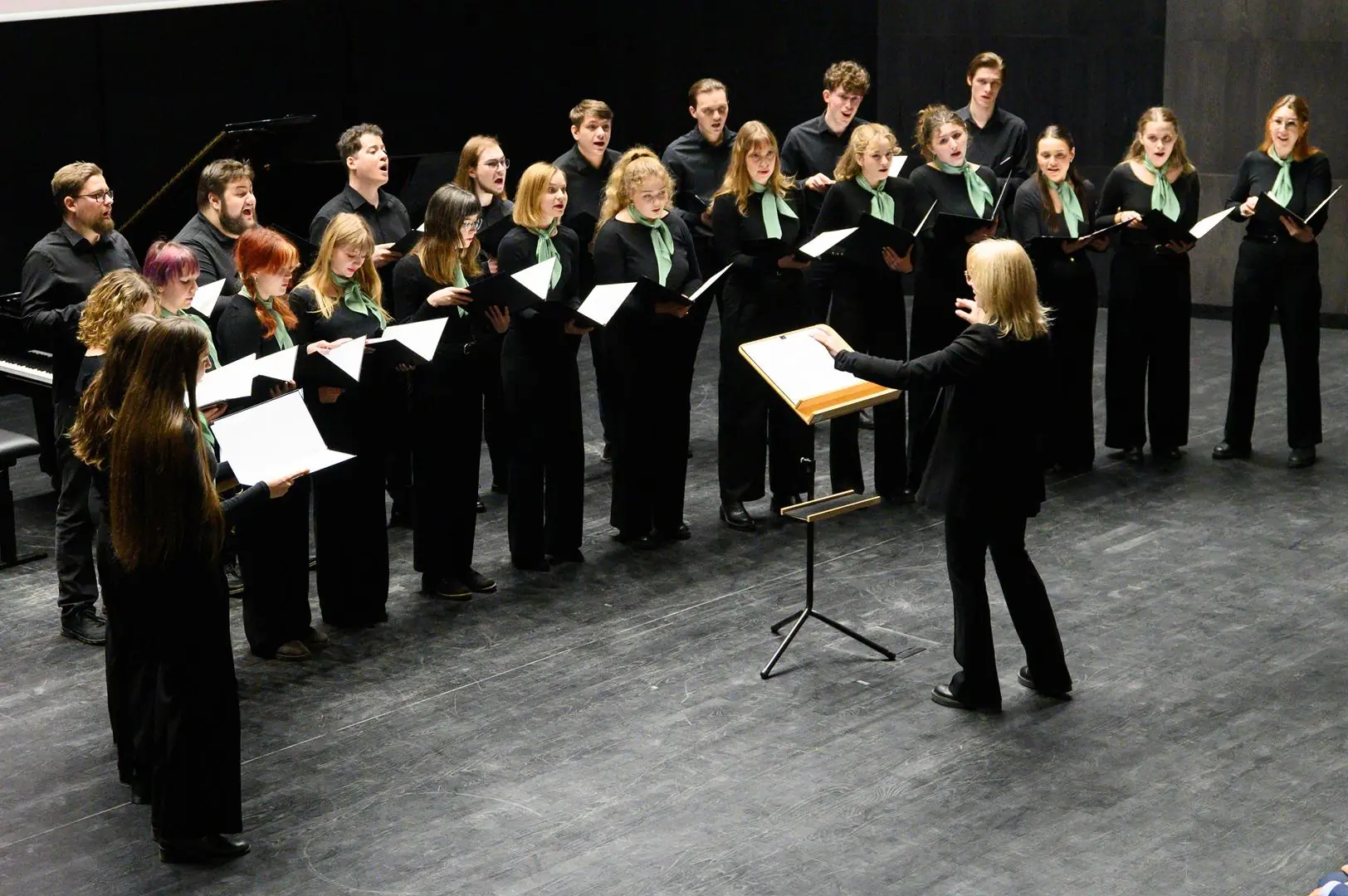 Chór stojący na scenie wykonuje utwór pod batutą dyrygentki.