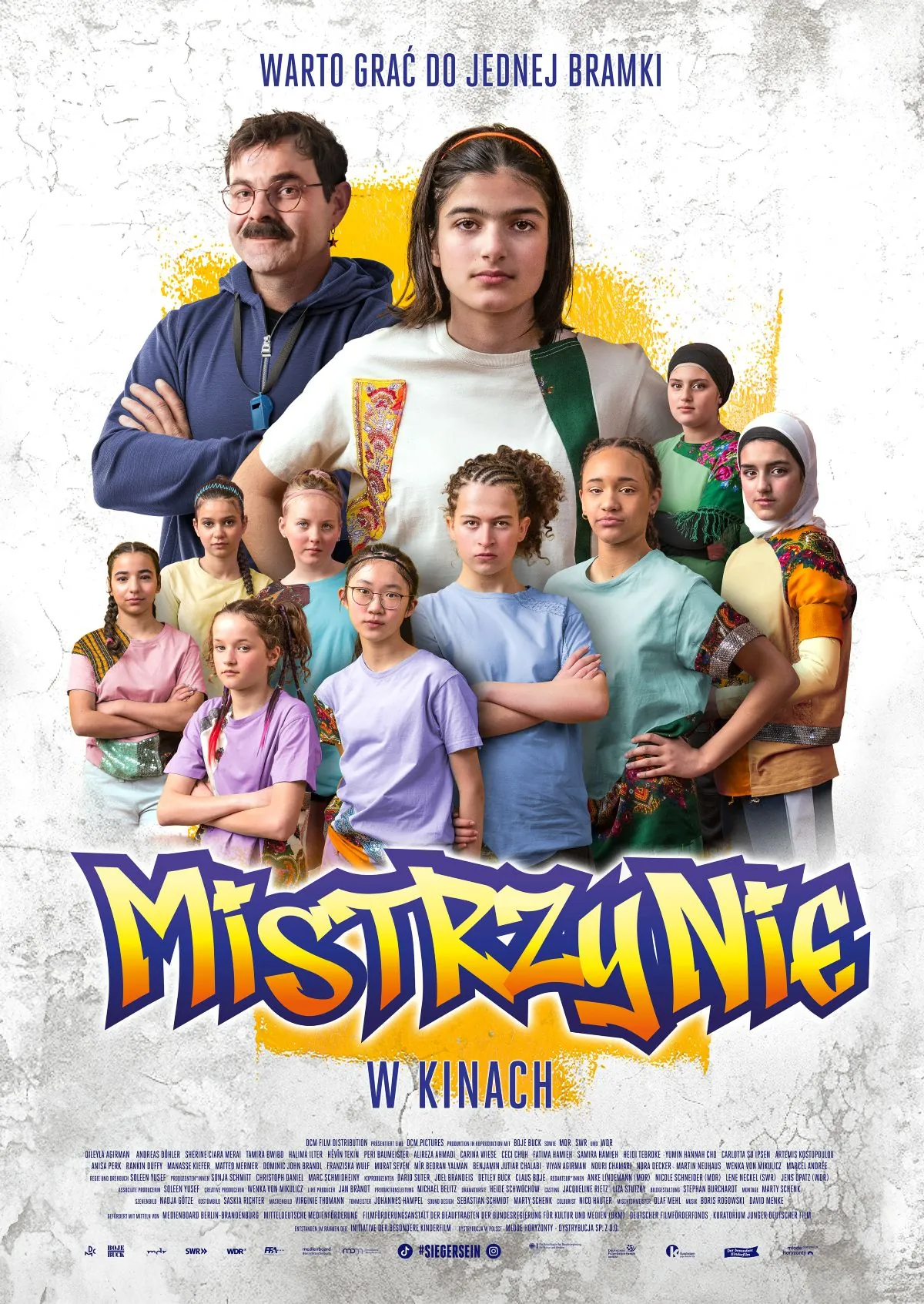 Plakat filmu „Mistrzynie”. W centrum stoi nastoletnia dziewczyna w jasnej koszulce z zielonym paskiem przewieszonym przez ramię, patrząca pewnie przed siebie. Za nią znajduje się mężczyzna w niebieskiej bluzie z gwizdkiem na szyi i skrzyżowanymi ramionami. Wokół nich stoi grupa dziewcząt o różnym pochodzeniu i wyglądzie, część w sportowych ubraniach, część w tradycyjnych strojach i chustach. U góry widnieje hasło „Warto grać do jednej bramki”, a u dołu duży tytuł „Mistrzynie” oraz napis „W kinach”.
